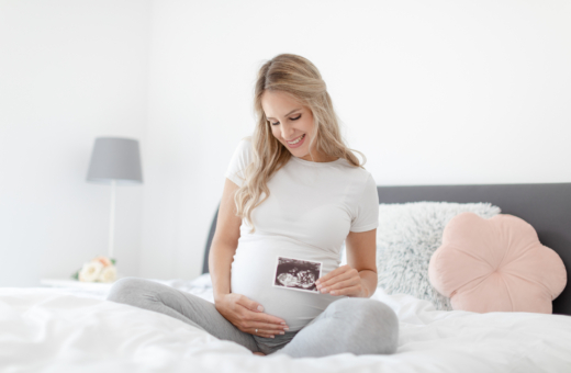 Rase õnnelik naine istumas voodis, ühe käega hoiab oma kõhtu, teises käes hoiab ultrahelipilti oma lootest.